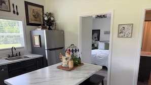Cocina con encimera de mármol, refrigerador y lavamanos. Hay un taburete de madera y una figurita decorativa de gallo sobre la encimera.