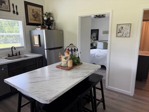 A kitchen with a marble countertop, a refrigerator, and a sink. There is a wooden stool and a decorative rooster figurine on the counter.