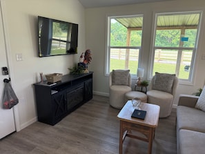 Ein Wohnzimmer mit einer schwarzen Unterhaltungsanlage, einem Fernseher, zwei Sesseln, einem Couchtisch und Blick auf eine grüne Landschaft durch große Fenster.