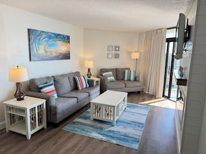 Ein Wohnzimmer mit grauem Sofa, gestreifteren Kissen, einem weißen Couchtisch, einem blau-weißen Teppich und einem Bild an der Wand.