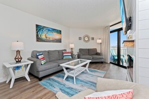 A modern living room with two grey sofas, a white coffee table, and a colorful rug. There are two side tables with lamps, a large painting of a wave, and a decorative mirror on the wall. - Amazing OceanView•14th-Fl• Spacious 3BR •Fireplace •Pool• Sleeps 10 (Myrtle Beach)