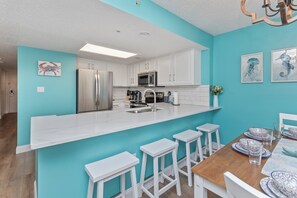 A modern kitchen with white cabinets, a stainless steel refrigerator, a microwave, and a sink. There are white bar stools and a wooden dining table with blue and white plates. - Amazing OceanView•14th-Fl• Spacious 3BR •Fireplace •Pool• Sleeps 10 (Myrtle Beach)
