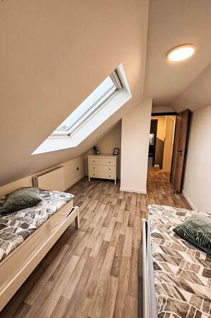 Ein Schlafzimmer mit schrägem Dach, einem Oberlicht, Holzboden, einem Bett, einer Kommode und einer Tür zu einem anderen Raum.