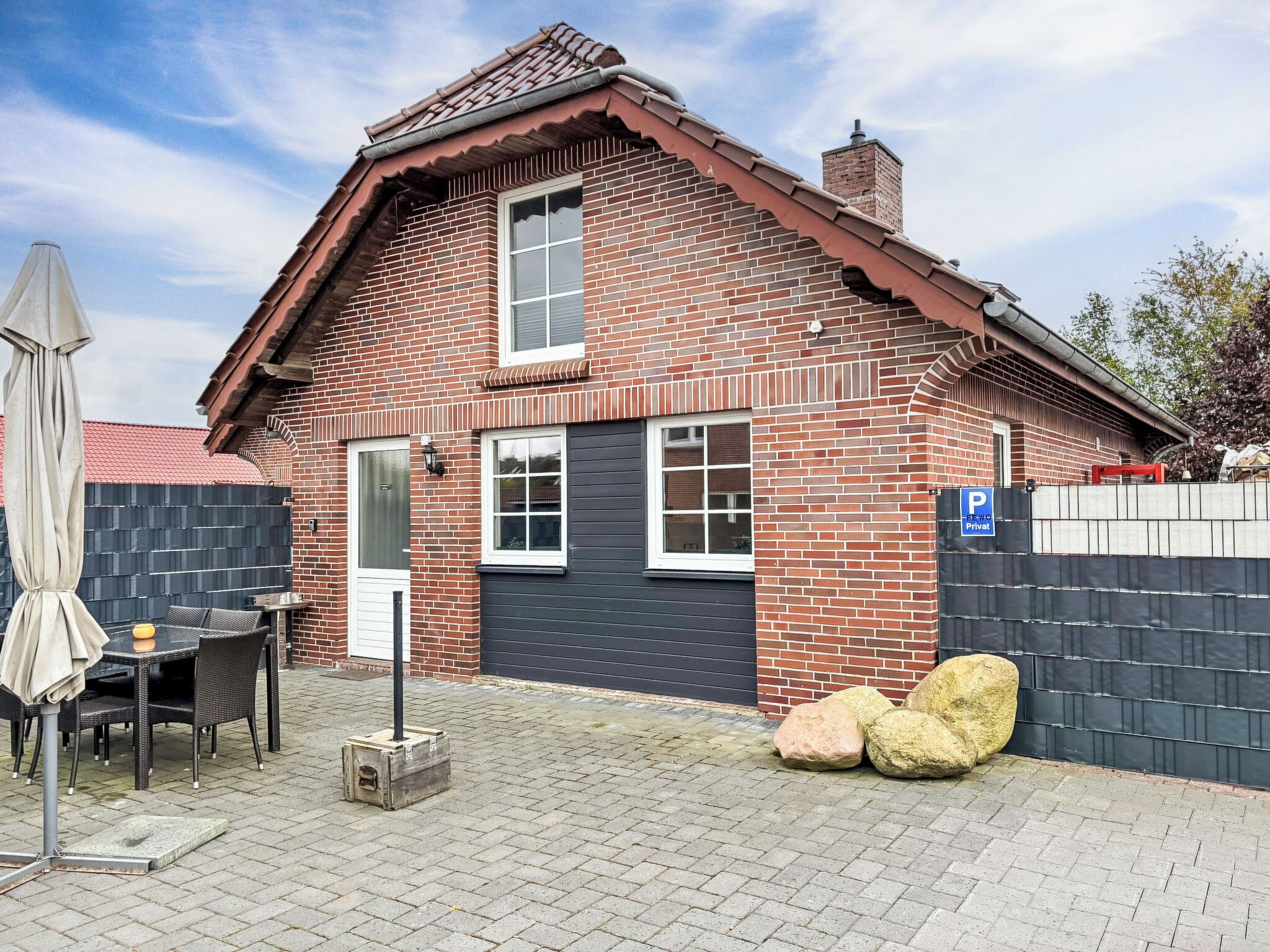 Une maison à deux étages, construite en briques, avec une porte de garage sombre, un petit patio doté d’une table et de chaises, et un panneau indiquant une place de stationnement privée.