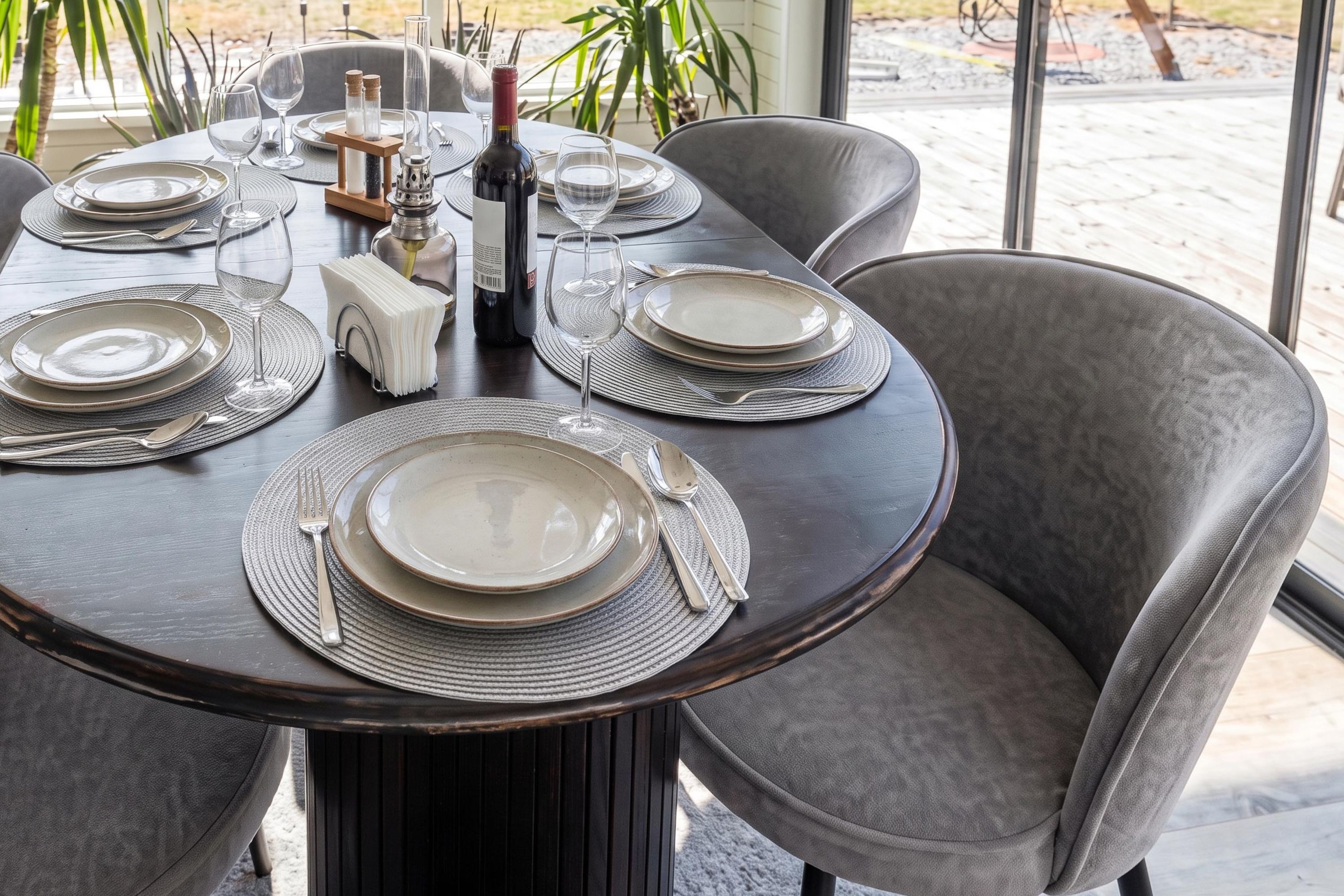 A dining table set for four with plates, cutlery, and a bottle of wine, surrounded by chairs.