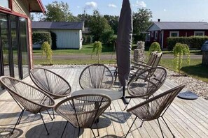 A deck with outdoor seating, a large umbrella, and a view of a red building and greenery. - Skummeslöv Family Escape B, Skummeslövstrand, Sweden (Skummeslövstrand)