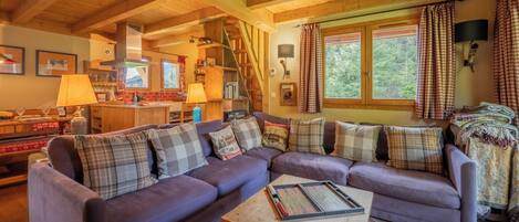 Una sala de estar acogedora con un sofá modular, vigas de madera, una cocina al fondo y un ventanal con cortinas.