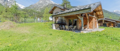 A wooden chalet with a sloped roof, a patio with outdoor seating, and a mountain backdrop.