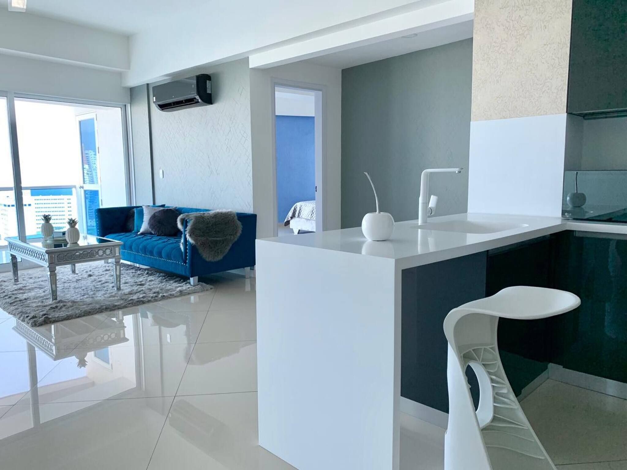 Uma sala de estar moderna com um sofá azul, uma mesa de centro branca e uma área de cozinha com bancada branca e uma cadeira branca.