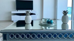 Een moderne woonkamer met een salontafel met glazen blad, witte decoratieve ananassculpturen en een flatscreen televisie op een witte kast.
