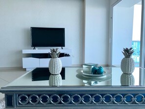 A modern living room with a glass-top coffee table, white decorative pineapple sculptures, and a flat-screen TV on a white cabinet. - Luxury 2 Bedrooms Water View Apartment (Cartagena)