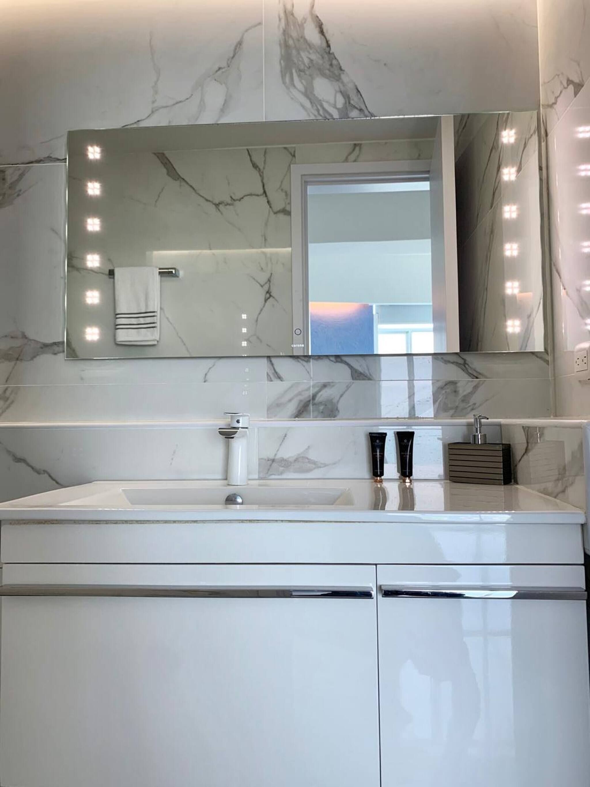 A bathroom with a marble vanity, a large mirror with lights, a towel rack, and a sink.