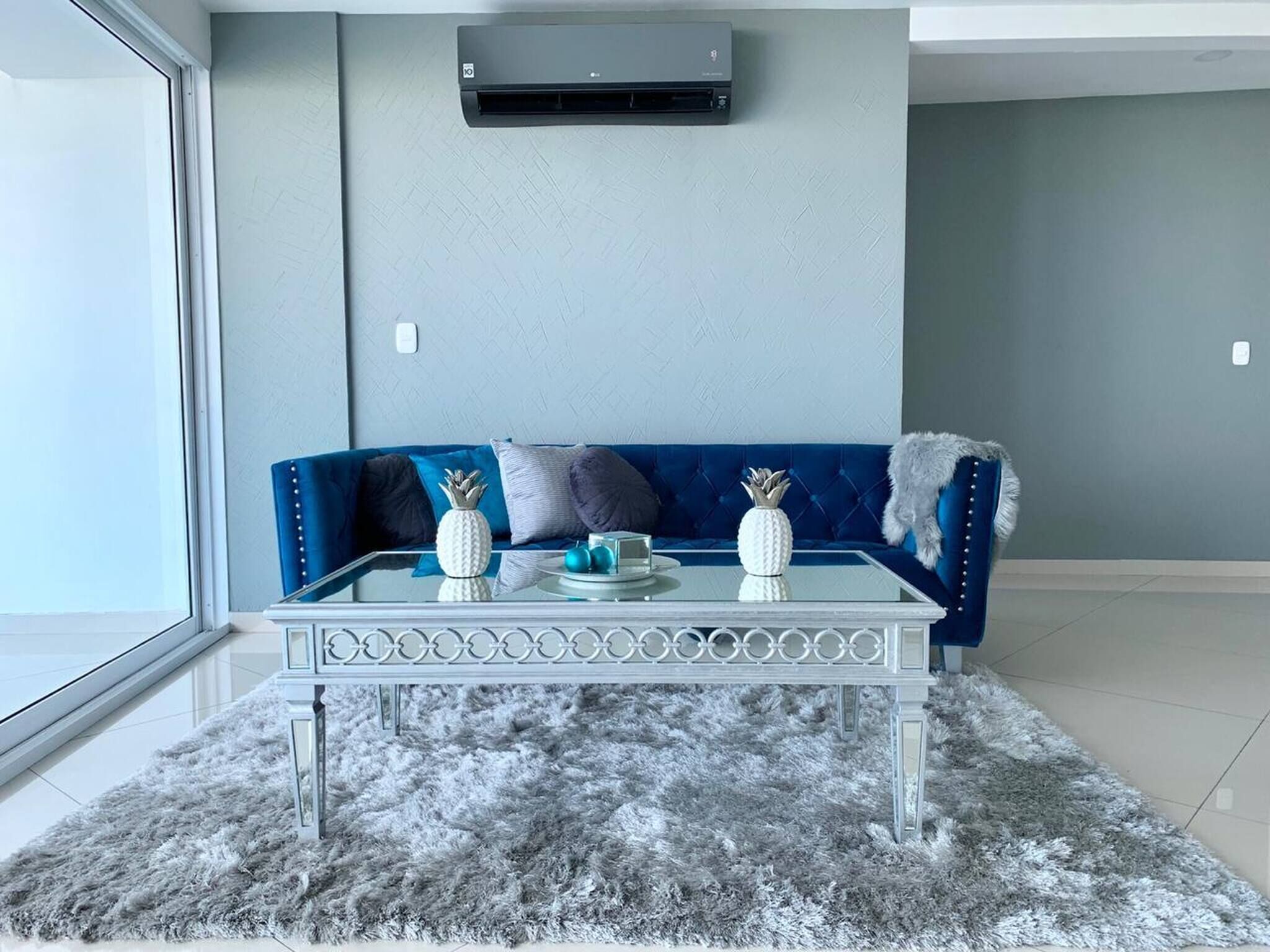 Una habitación con un sofá azul, una mesa de centro de vidrio y un tapete gris y mullido.