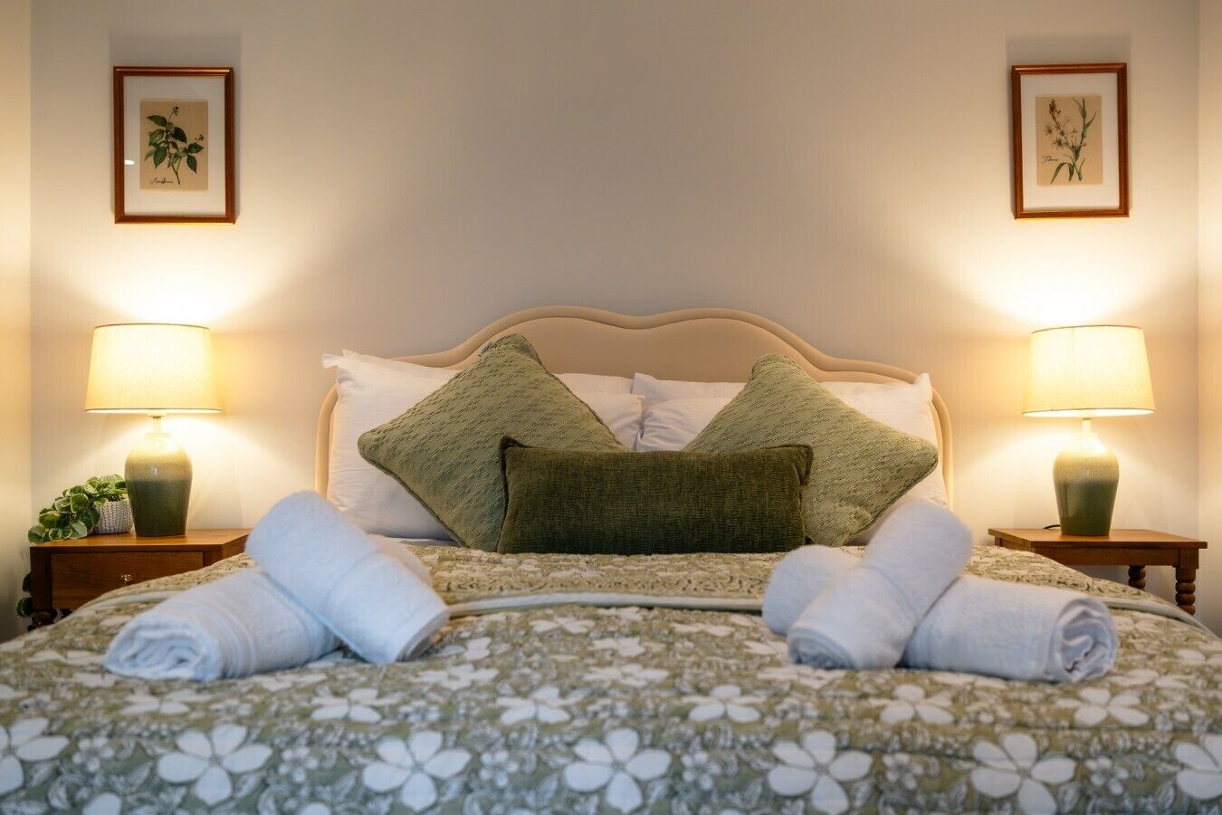 Una cama bien tendida con una colcha estampada con flores, dos almohadas verdes y dos lámparas de noche.