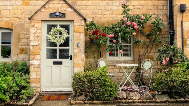 A stone cottage with a white door, a small porch with a chair, and a sign reading 'Little Cottage.'