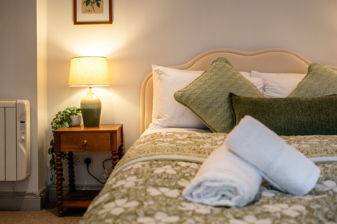 Un letto rifatto con cura, con una coperta a motivi floreali, un cuscino verde e un asciugamano bianco arrotolato. Su un comodino in legno c'è una lampada da letto e una pianta.