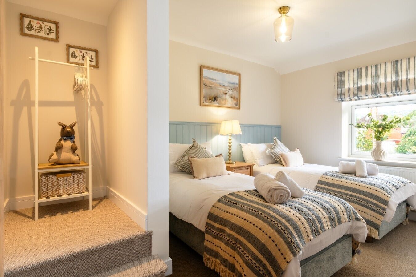 A bedroom with a bed, a nightstand, a lamp, a window with curtains, and a wall-mounted shelf with a stuffed toy.