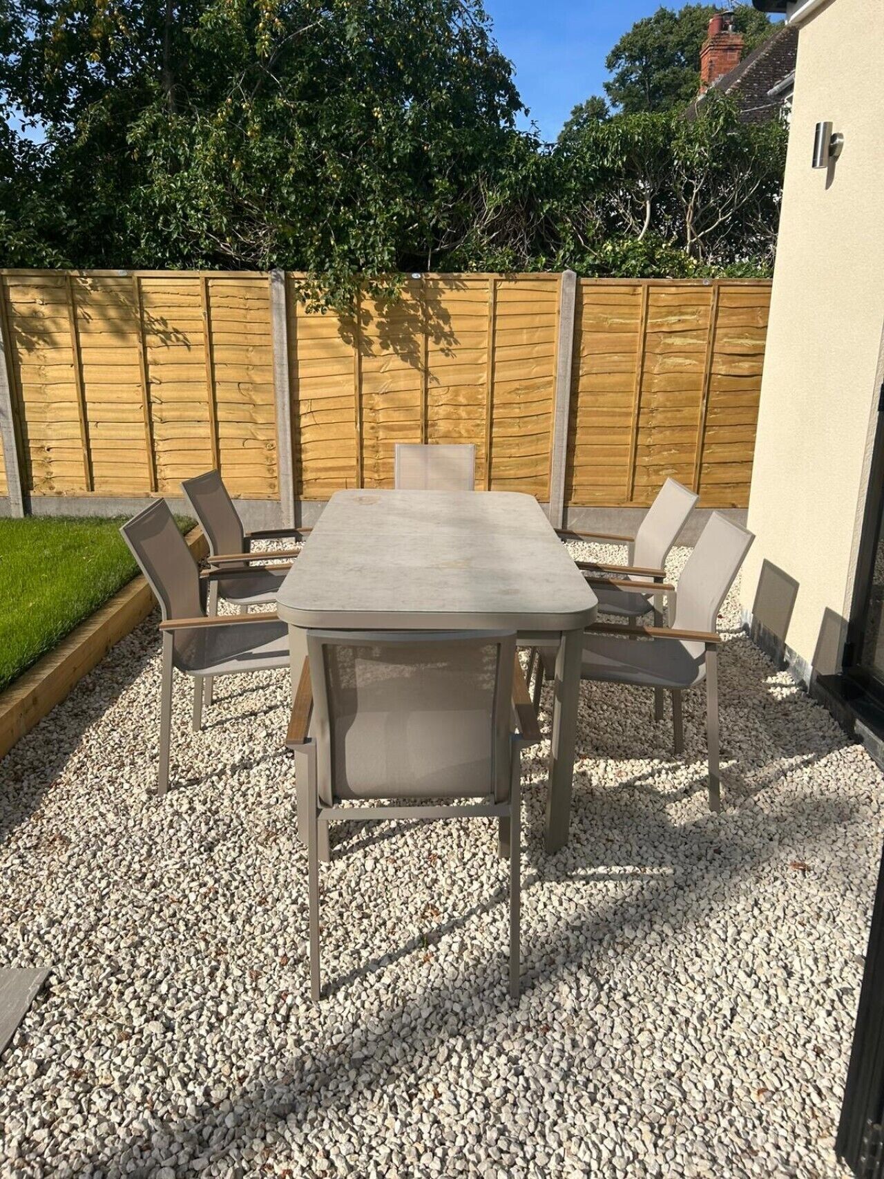 A patio with a rectangular table and six chairs, surrounded by a wooden fence and gravel ground.