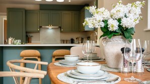 Een eetruimte met een houten tafel gedekt voor vier personen, omringd door stoelen, met een vaas witte bloemen op de tafel.