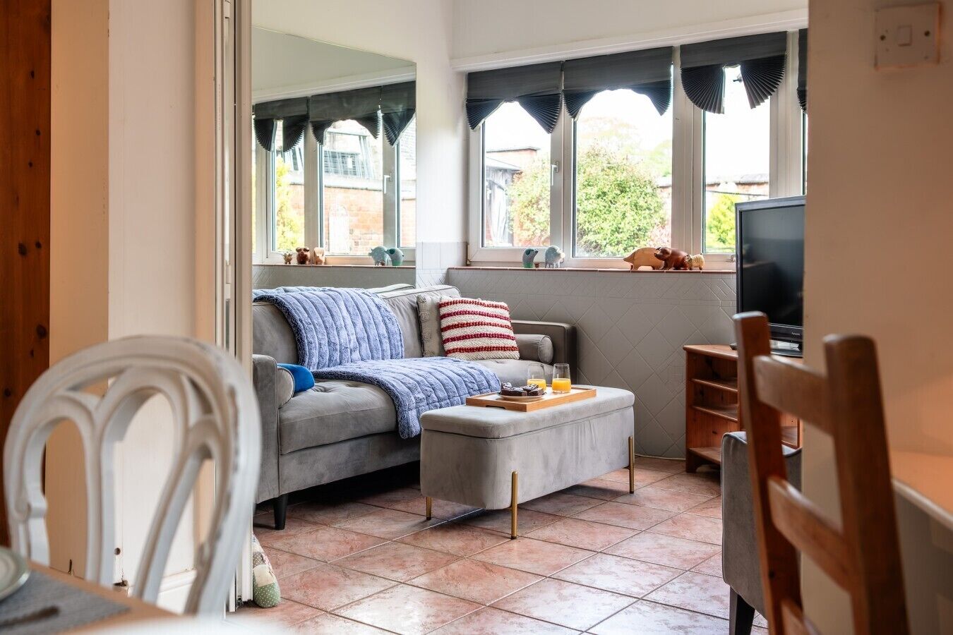 Un salon chaleureux avec un canapé, un pouf et un téléviseur, doté de grandes fenêtres et d’un sol carrelé.