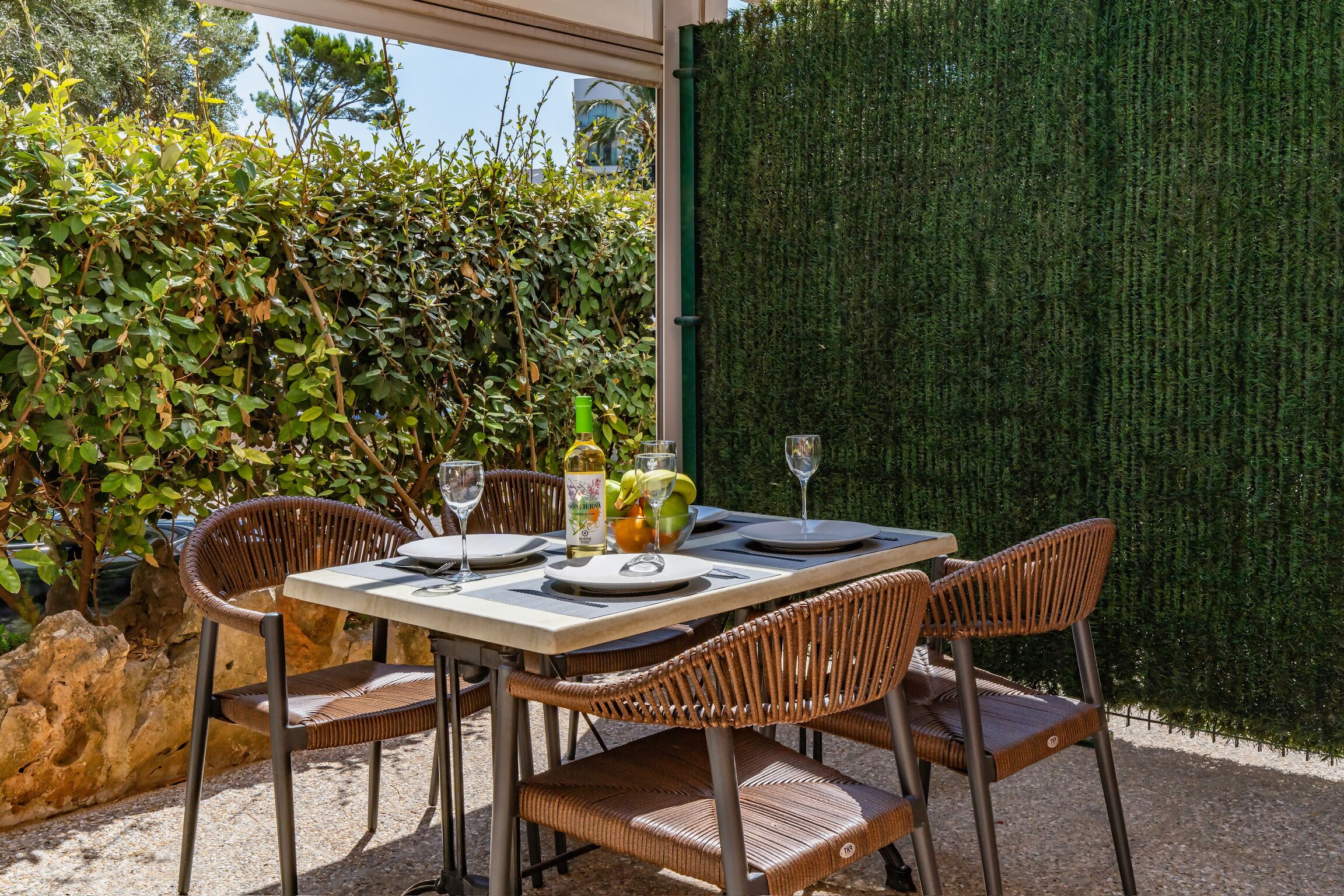 Un espace de restauration en plein air, avec des chaises en osier et une table dressée pour deux personnes, entouré de verdure et d’une haie.