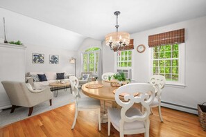 A dining area with a round table, white chairs, a chandelier, and large windows with views of greenery. - Sunset Pines Cape Cod retreat (East Sandwich)