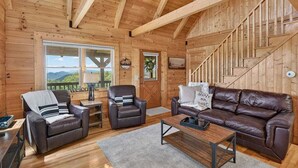 Interior de una cabaña de madera con sofá, dos sillones, una mesa de centro, una escalera y un ventanal con vistas.
