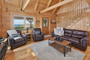 Ein Holzblockhaus-Innenraum mit einer Couch, zwei Sesseln, einem Couchtisch, einer Treppe und einem großen Fenster mit Blick.