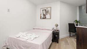 A bedroom with a bed, a bedside table with a lamp, a chair, a desk with a plant, and a wall-mounted picture.