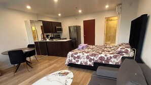 A modern living room with a sofa, a dining table with chairs, a kitchen area with a refrigerator and microwave, and a bedroom with a bed and a bathroom visible through a glass door.