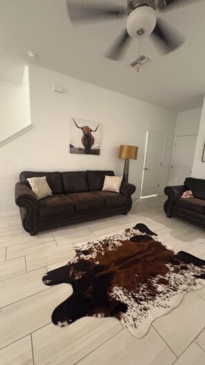 Een woonkamer met een bruine leren bank, een kleden van runderhuid, een plafondventilator en een deur op de achtergrond.