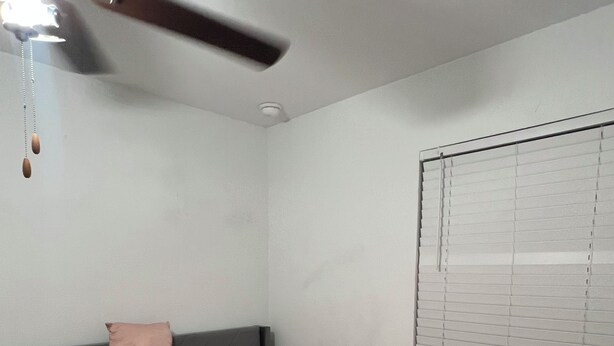 A bedroom with a bed, a ceiling fan, a window with blinds, and a wall-mounted light fixture.