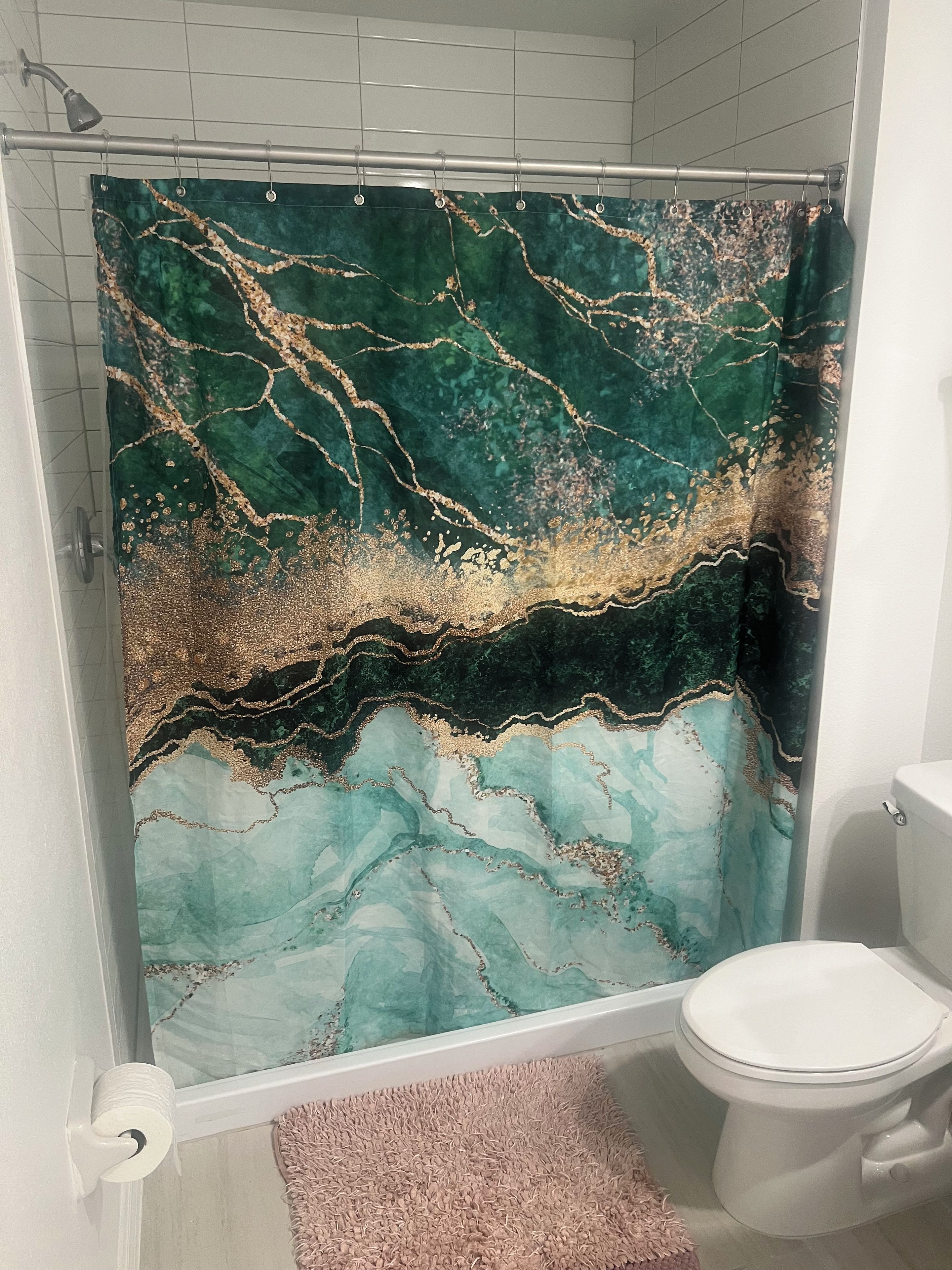 Une salle de bain avec un rideau de douche orné d’un motif rappelant le marbre, des toilettes et un tapis rose.