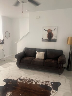 Ett vardagsrum med en brun lädersoffa, en matta av kohud, en tavla med en bild av en tjur som hänger på väggen, en takfläkt och en trappa.