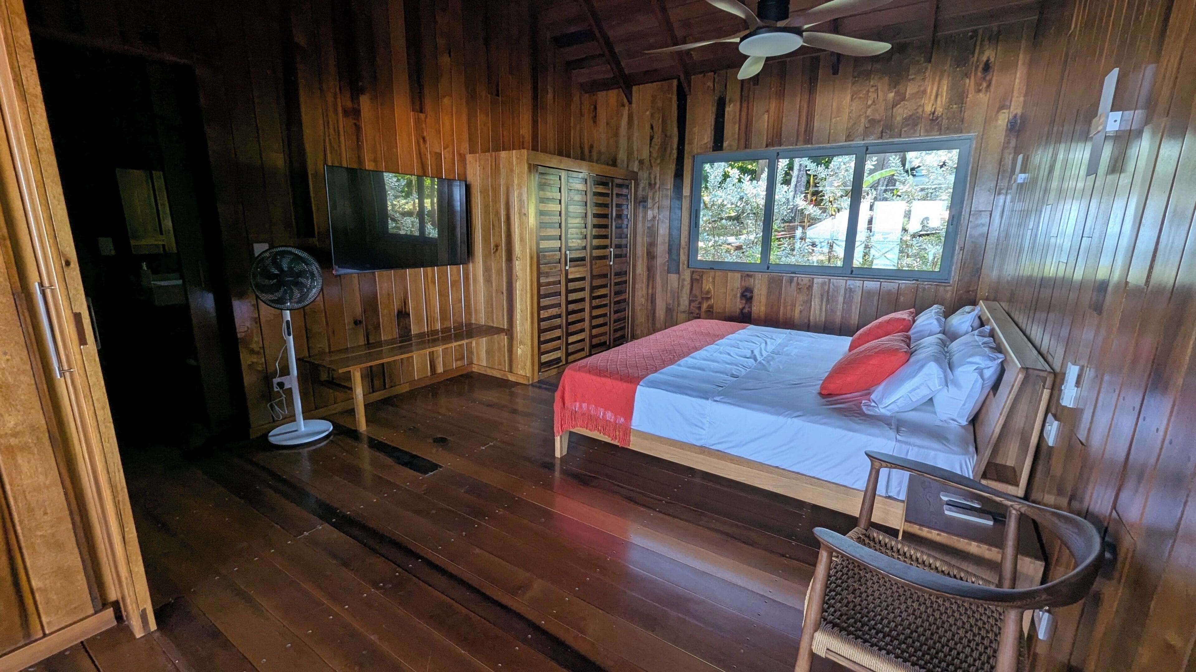Ein Zimmer mit Holzboden, einem Bett, einem Fernseher, einem Ventilator und einem Fenster.