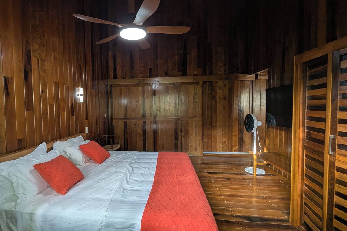 A wooden room with a bed, ceiling fan, and a fan on the floor.