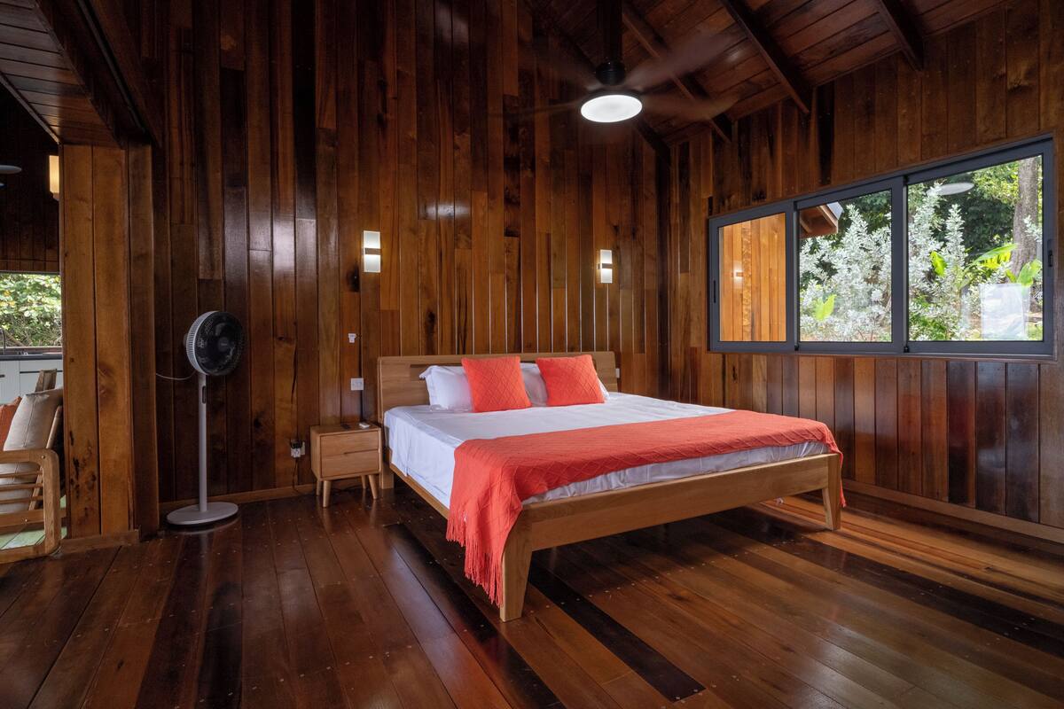 A wooden-paneled room with a bed, a nightstand, a floor fan, and a window with a view of greenery.