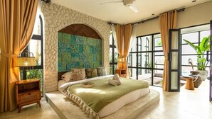 Une chambre avec une arche en pierre, un lit recouvert d’une couverture verte, une table de chevet et une vue dégagée sur l’extérieur grâce à de grandes fenêtres.