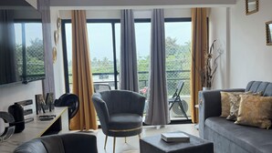 Uma sala de estar moderna com sofá, poltrona e mesa de centro. Há um ventilador de teto, janelas amplas com cortinas e vista para o verde lá fora.