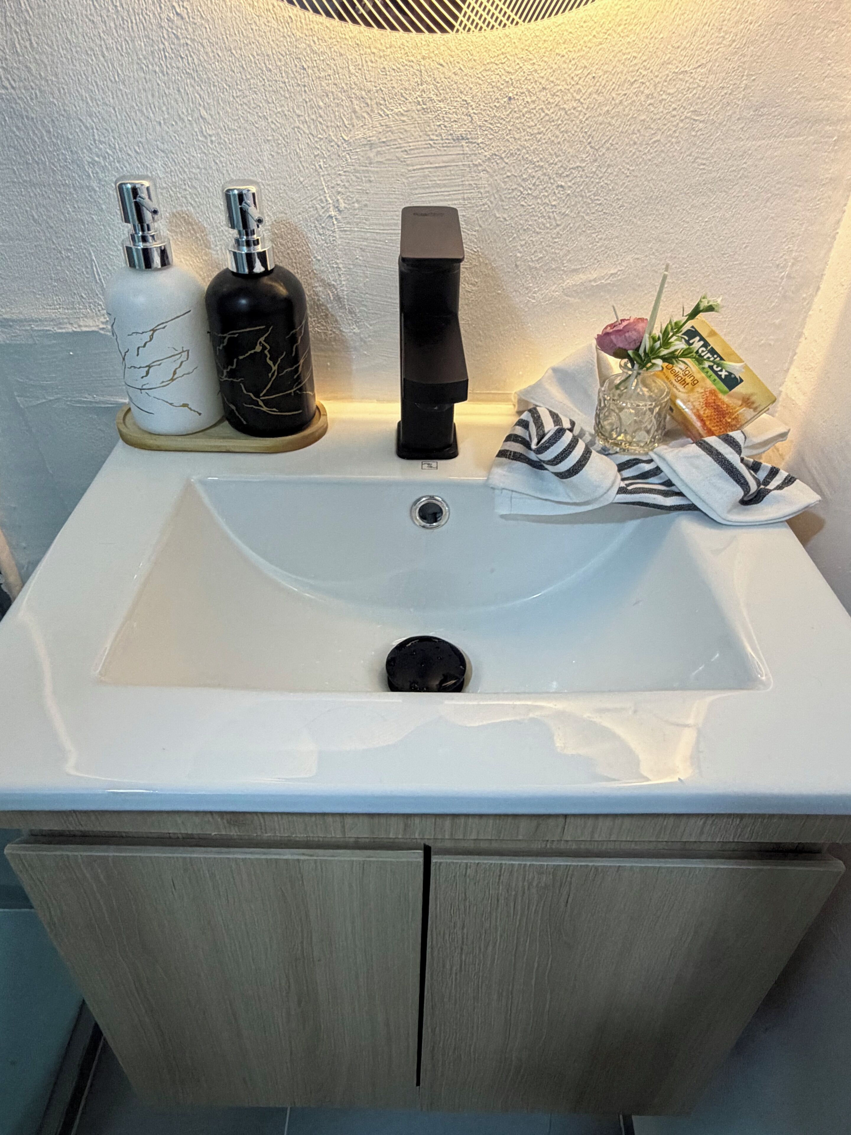 Een badkamer met een witte wastafel, twee zeepdispensers, een zwarte kraan, een glazen vaas met een roos en een handdoek.