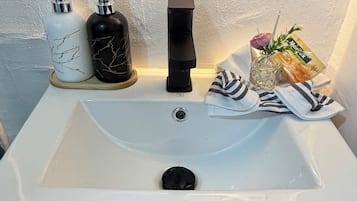 Een badkamer met een witte wastafel, twee zeepdispensers, een zwarte kraan, een glazen vaas met een roos en een handdoek.
