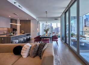 Una cocina moderna con taburetes de bar, un comedor con mesa redonda y sillas, y un ventanal con vistas a la ciudad.