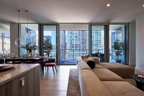 Una sala de estar moderna con sofá, mesa de comedor y sillas, ventanas amplias con vistas a la ciudad, y lámparas colgantes.