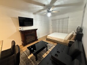 Ein modernes Wohnzimmer mit einem Flachbildfernseher, einem dunklen Holzschrank, einem schwarzen Couchtisch, einem schwarzen Sofa, einem Deckenventilator und einem Fenster mit Jalousien.