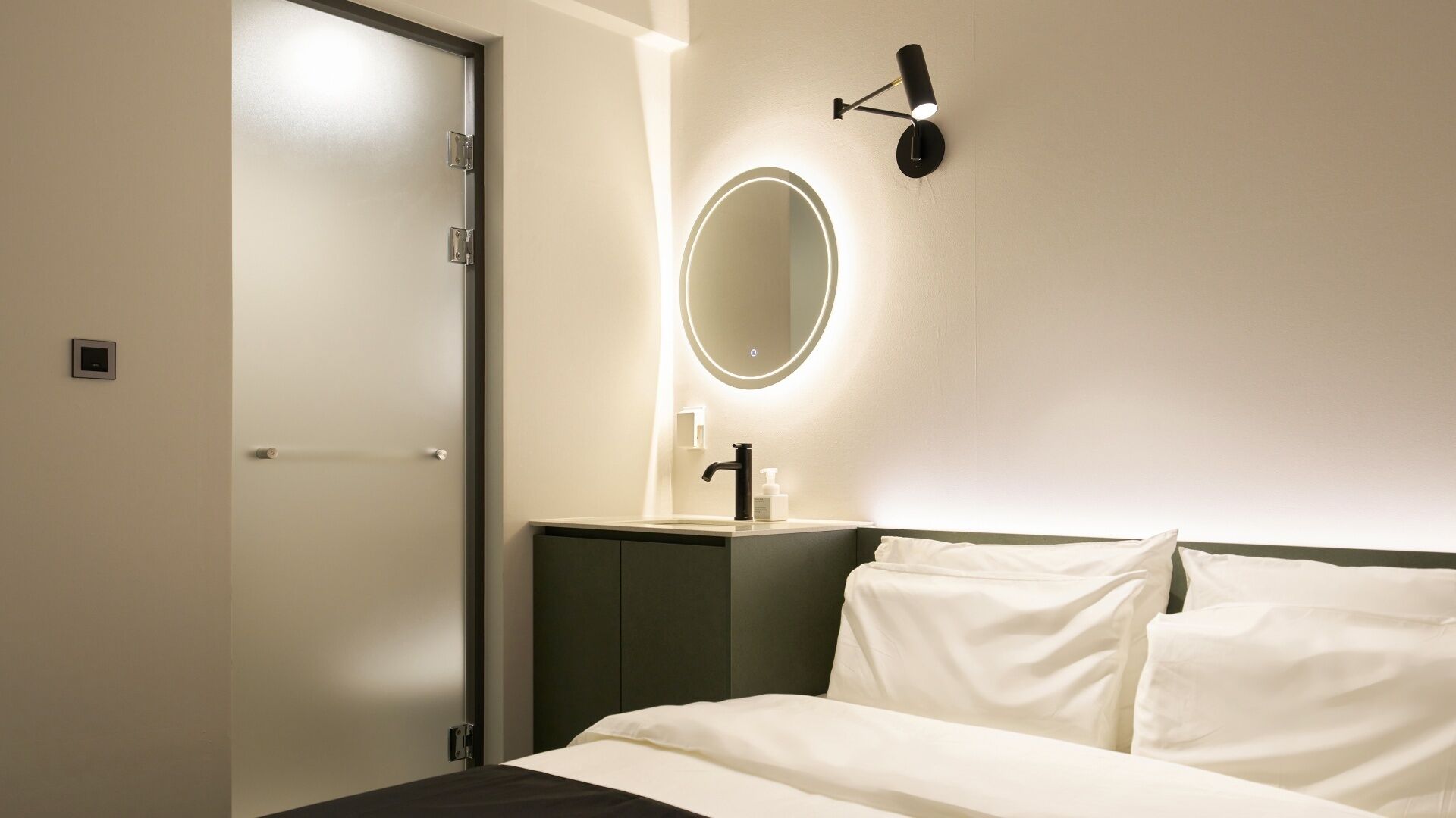 A hotel room with a bed, a bedside table, a mirror, and a wall-mounted light fixture.