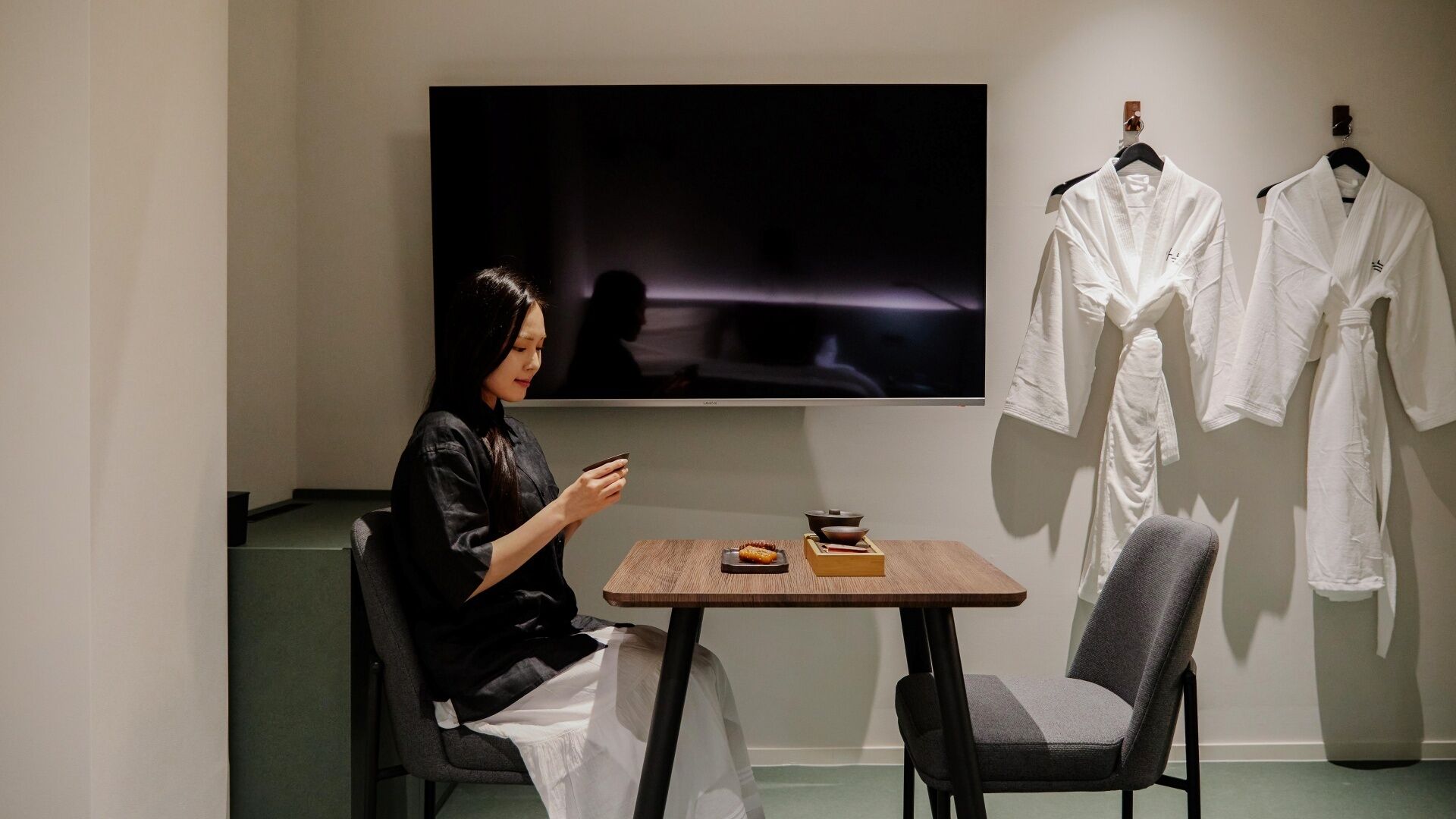 A person seated at a table with a television, a chair, and a table with a bowl and a cup.