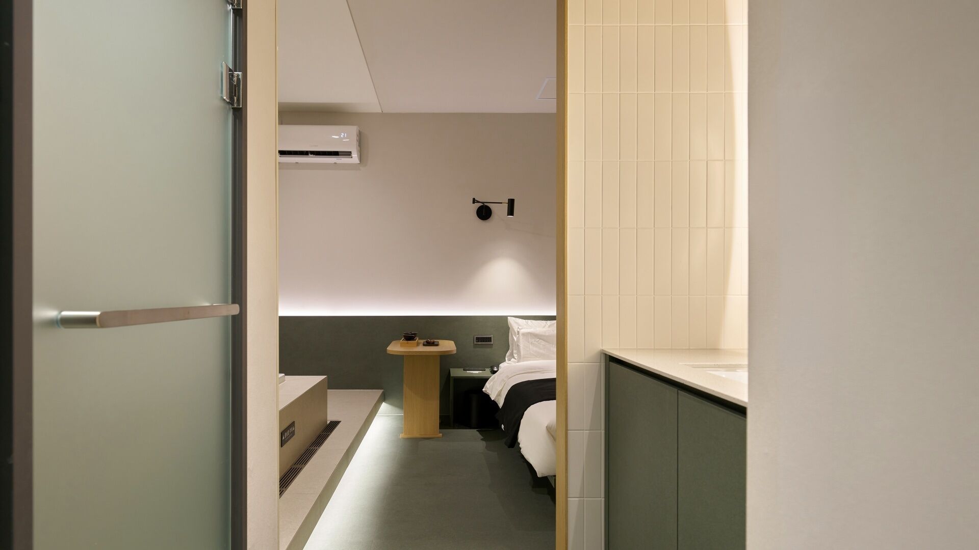 Un pasillo estrecho que conduce a un dormitorio con cama, mesita de noche y aire acondicionado instalado en la pared.