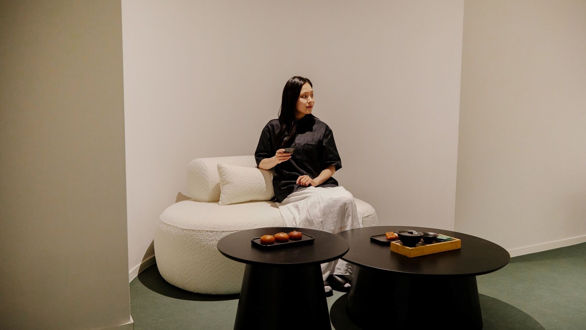 A person seated on a white, round, cushioned chair in a minimalist room with two black tables, one with a tray of food.