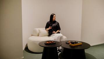 A person seated on a white, round, cushioned chair in a minimalist room with two black tables, one with a tray of food.