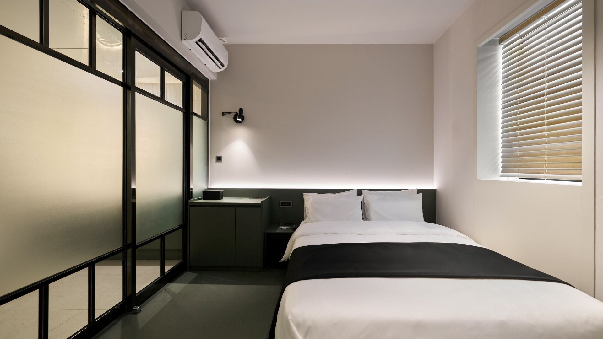 Habitación de hotel pequeña con cama, mesita de noche y ventana con persianas.